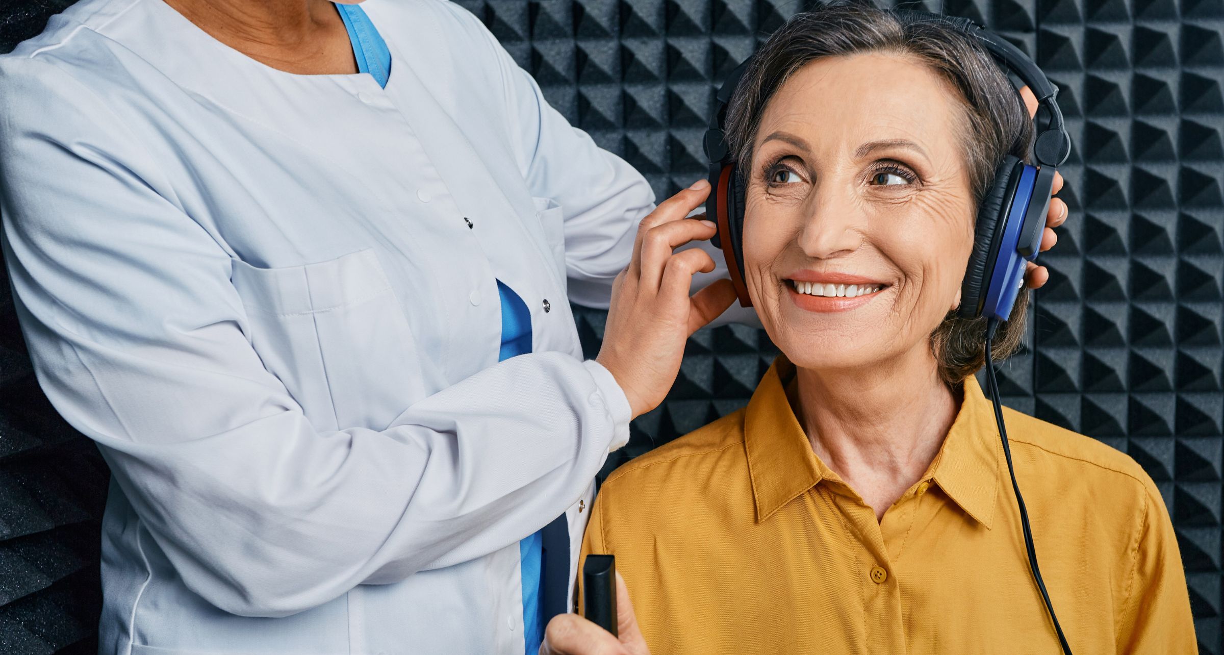 Signora che sorride mentre sta eseguendo degli esami audiometrici all'interno della cabina con le cuffie alle orecchie messe dall'audioprotesista