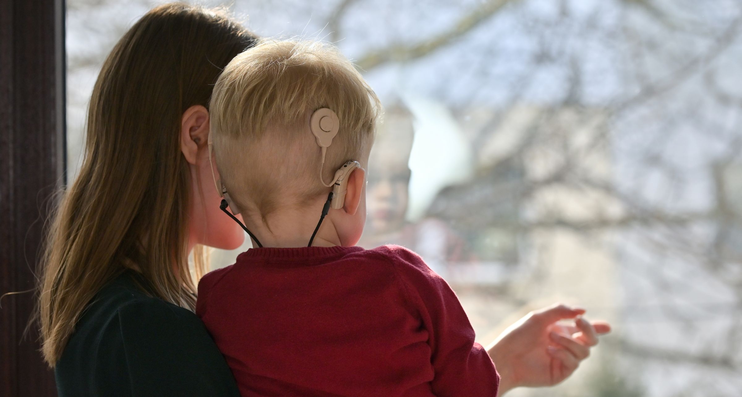 Mamma tiene in braccio bambino con impianti cocleari e entrambi di spalle guardano fuori dalla finestra